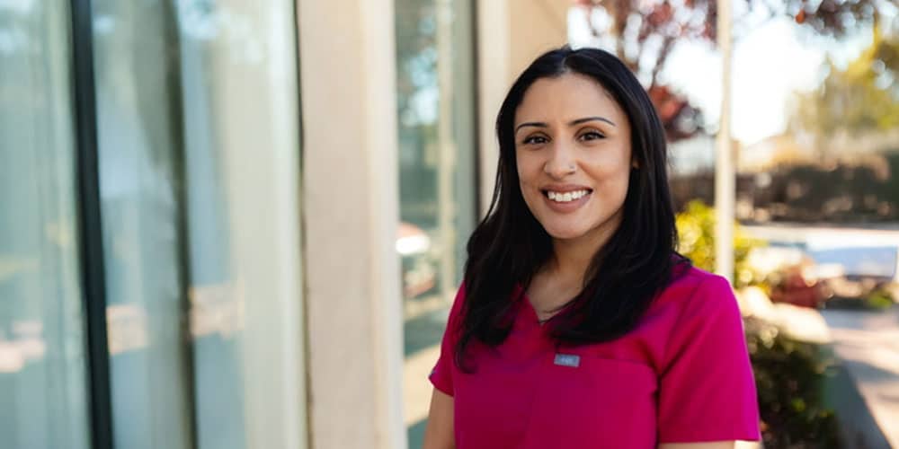 A woman with long dark hair, wearing a bright pink scrub top, stands outside near a building with large windows, smiling at the camera on a sunny day.