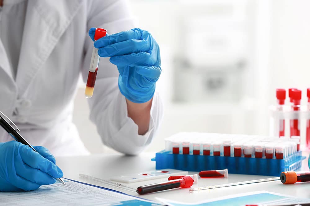 A person wearing a white lab coat and blue gloves examines a blood sample in a test tube, surrounded by other blood-filled tubes in a rack and laboratory equipment on a desk.
