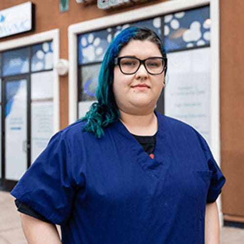A person wearing glasses and a blue scrub top stands in front of a building with pet-related signage. Their hair, partially dyed blue, hints at their vibrant personality as a dedicated veterinarian. They look at the camera with a neutral expression, embodying the calm demeanor of an experienced vet.