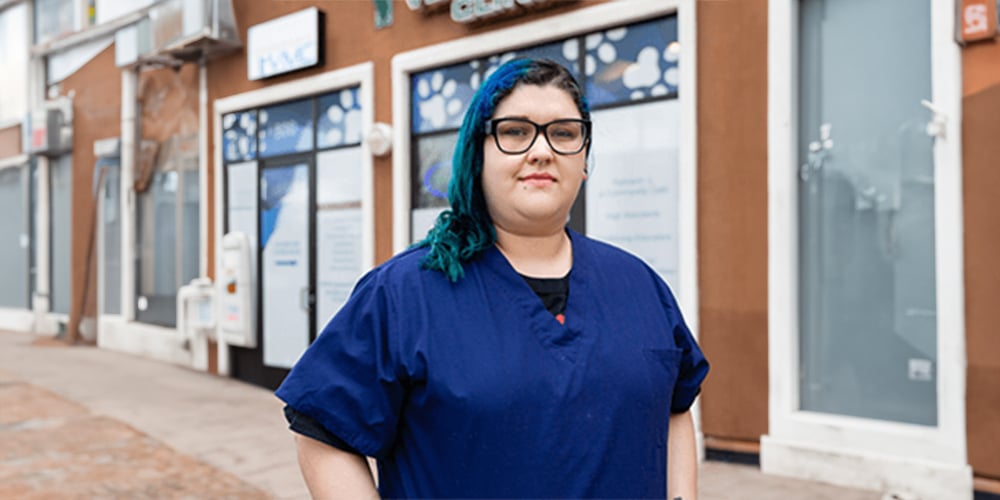 A person with glasses and blue-tinted hair wearing a dark blue outfit stands confidently outside a veterinary clinic, the signage hinting at their role as a vet. The background reveals a walkway lined with storefronts and large windows reflecting the busy street scene.