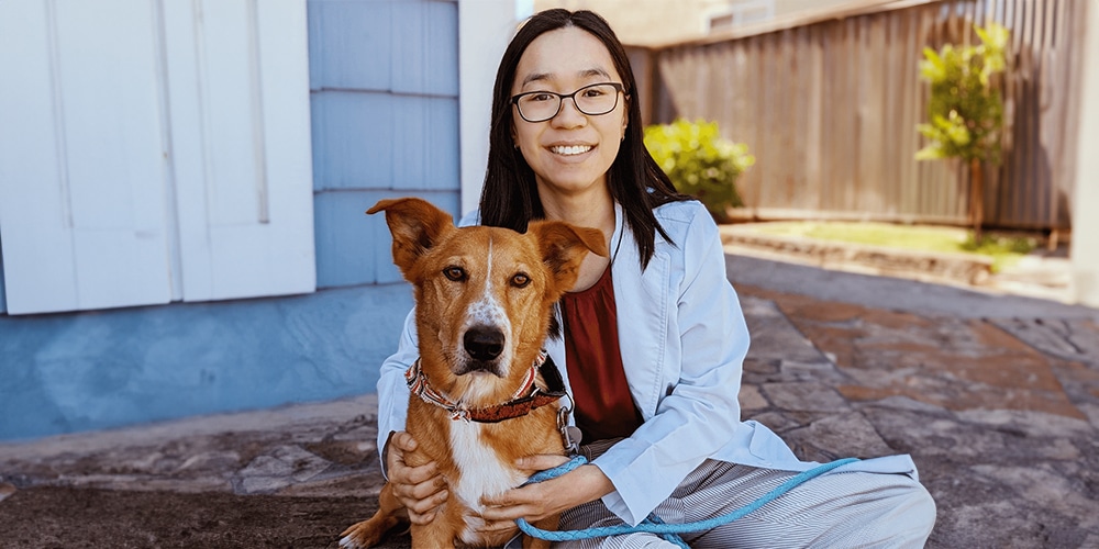 A person with glasses and long hair sits on the ground, smiling, like a caring veterinarian, while holding a mixed-breed dog wearing a red collar and leash. They are outside near a blue and white building, with a wooden fence in the background.