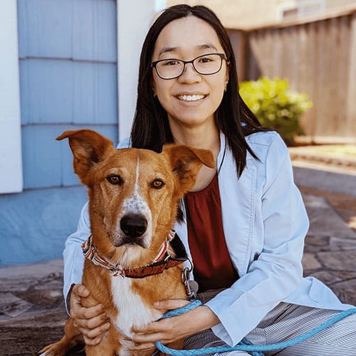A woman with glasses, wearing a light blue jacket and red shirt, sits on a stone path outside. She smiles while holding a brown and white dog on a leash, embodying the serene connection of a veterinarian to her beloved animal friends. A wooden fence and greenery are in the background.