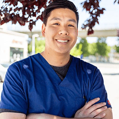 A veterinarian wearing blue scrubs stands outdoors with arms crossed, smiling. A tree with red leaves and a clear sky form the backdrop, suggesting a sunny day.