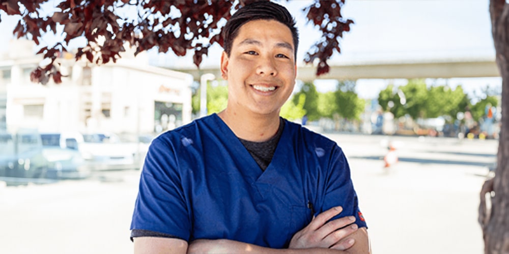A veterinarian in blue scrubs smiles while standing outdoors. They are under a tree with red leaves, with a street and buildings in the background. It's a sunny day.