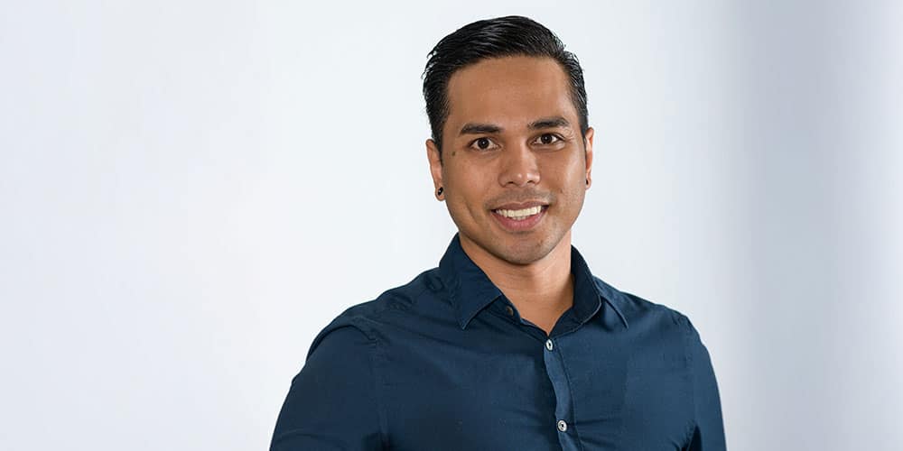 A man with short dark hair and a neatly groomed beard, wearing a dark blue button-up shirt, stands smiling against a plain, light background.