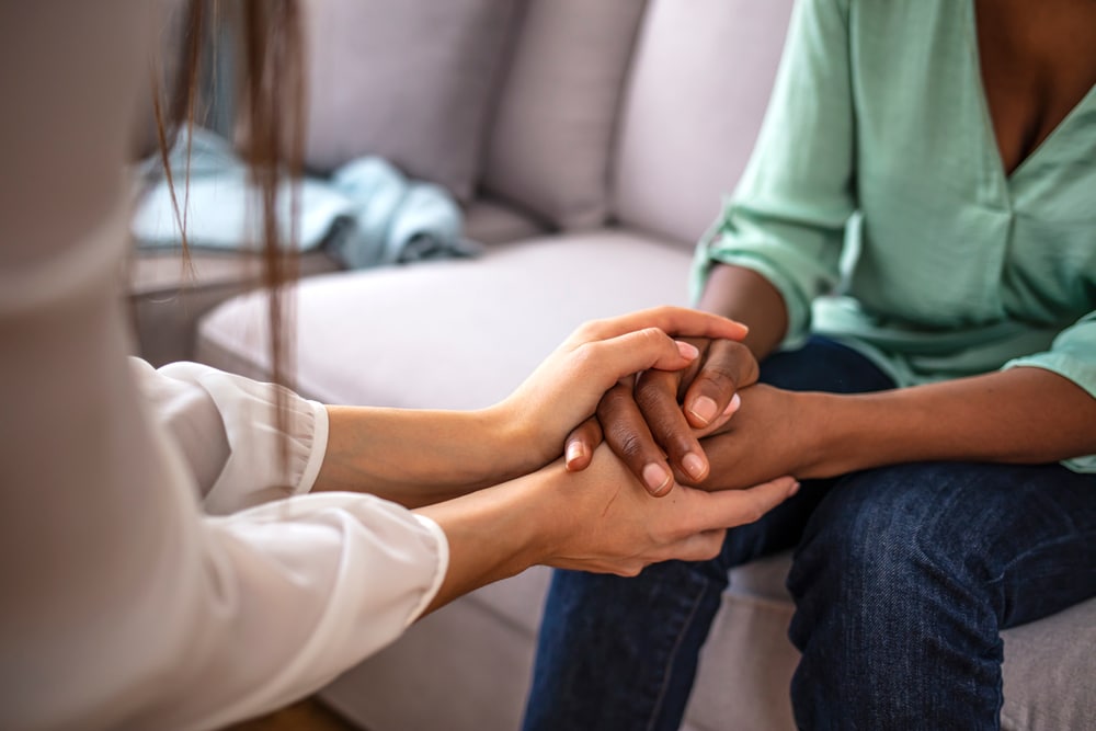 Two people sit close on a couch, where one, possibly a vet, gently holds the hands of the other, offering comfort or support. The setting appears to be a cozy, casual indoor space.